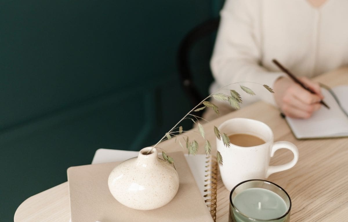 Interior designer writing notes about her ideal client in a notebook beside a cup of coffee and a ceramic vase