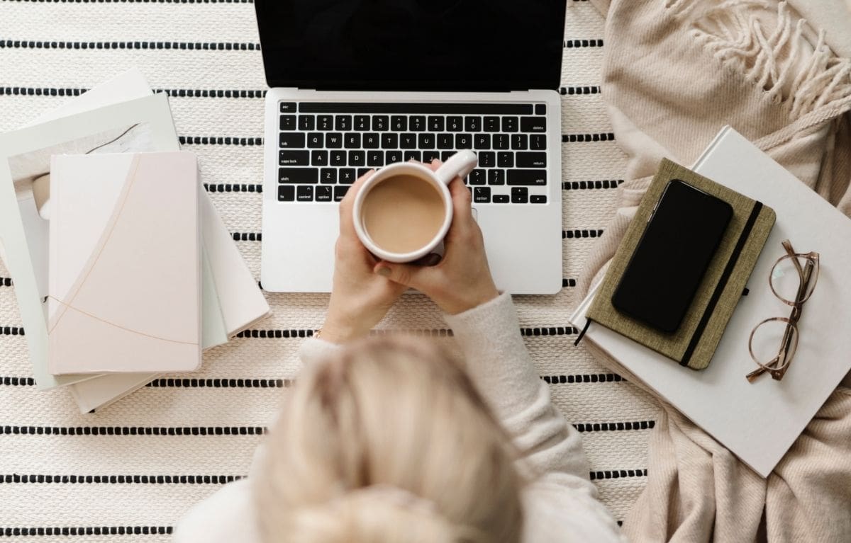 Interior designer holding a cup of coffee over her laptop with notebooks and glasses nearby, preparing an interior design website checklist